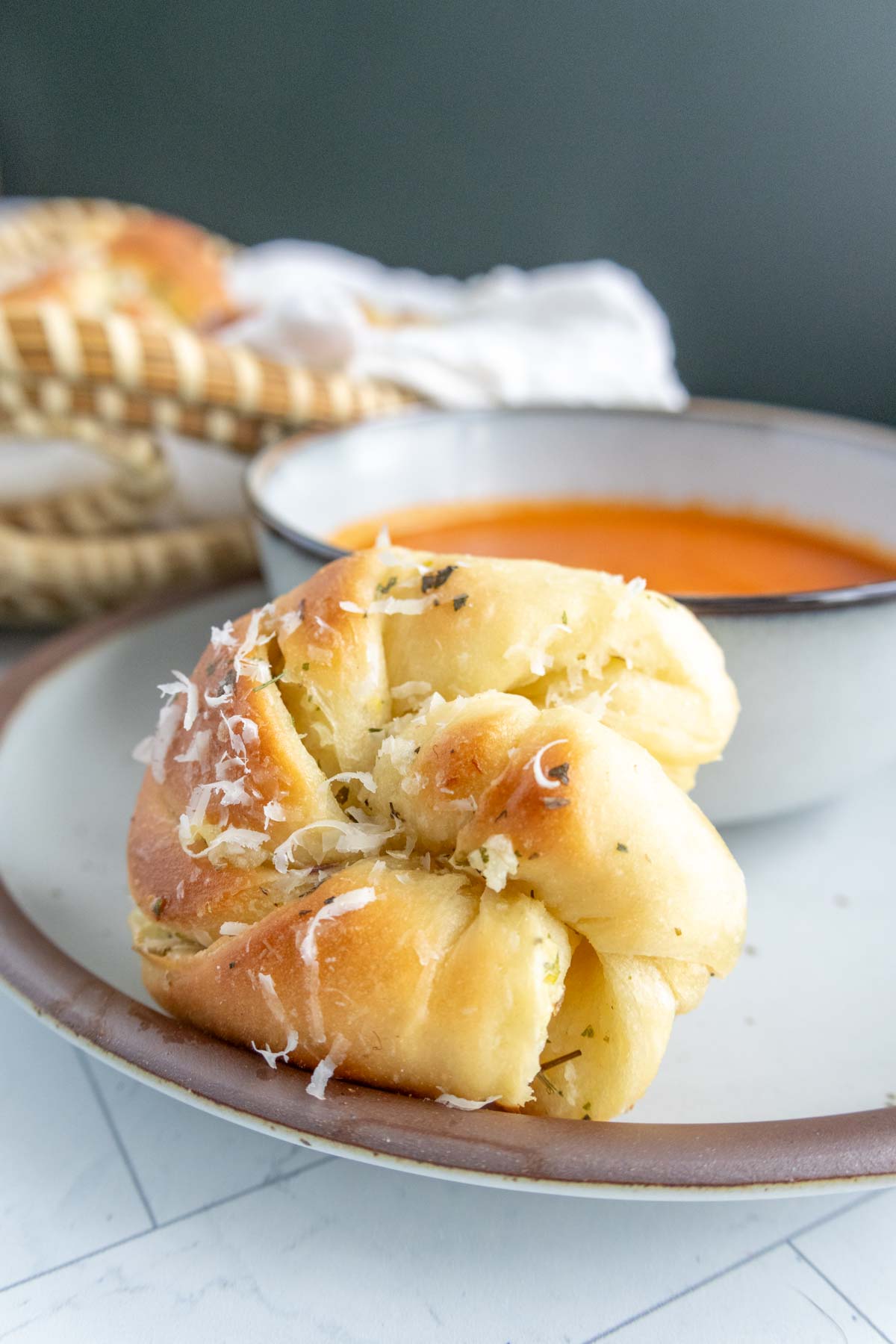 A garlic knot sprinkled with cheese on a plate next to a bowl of tomato soup.