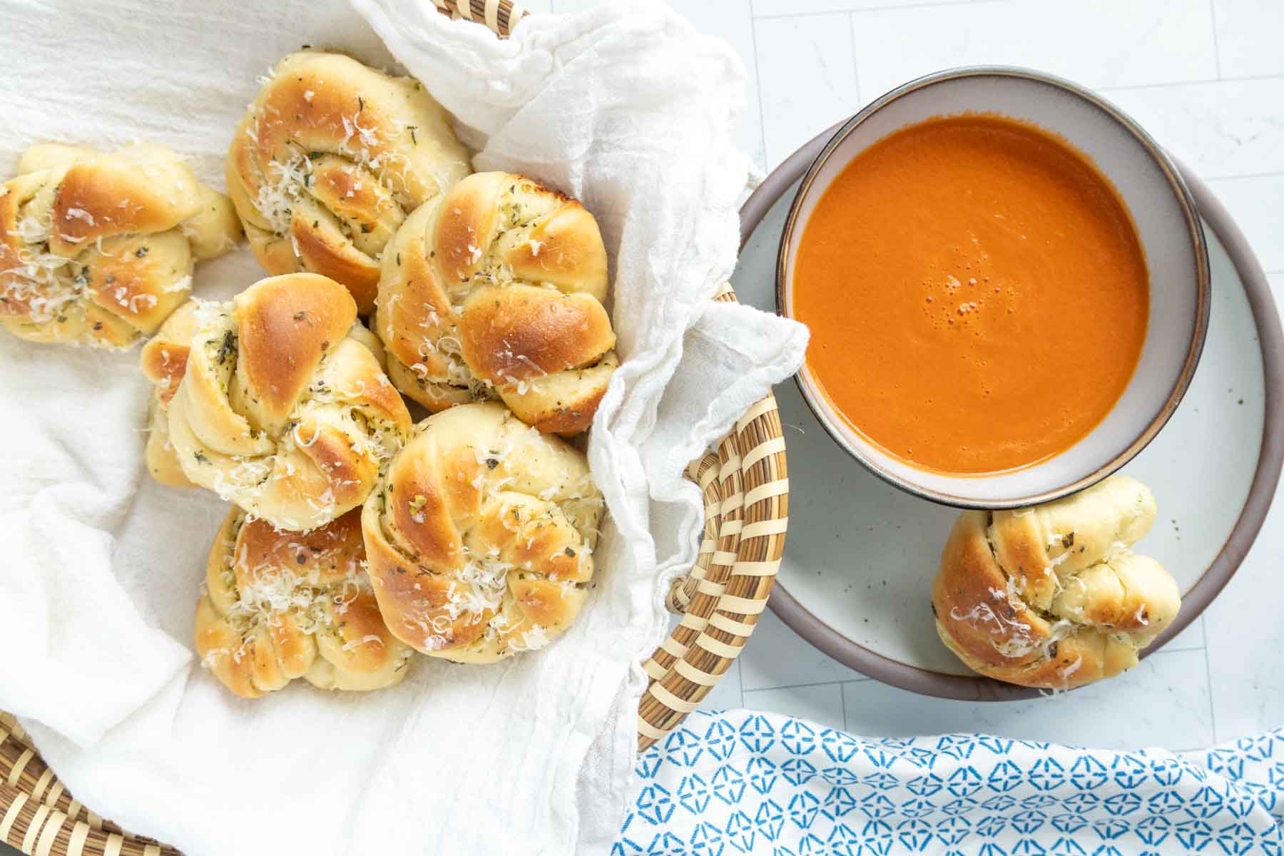 Basket of garlic knots with grated cheese alongside a bowl of tomato soup on a plate with an extra garlic knot.