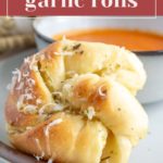 A close-up of soft garlic rolls on a plate, garnished with grated cheese. In the background, there's a bowl of orange soup.