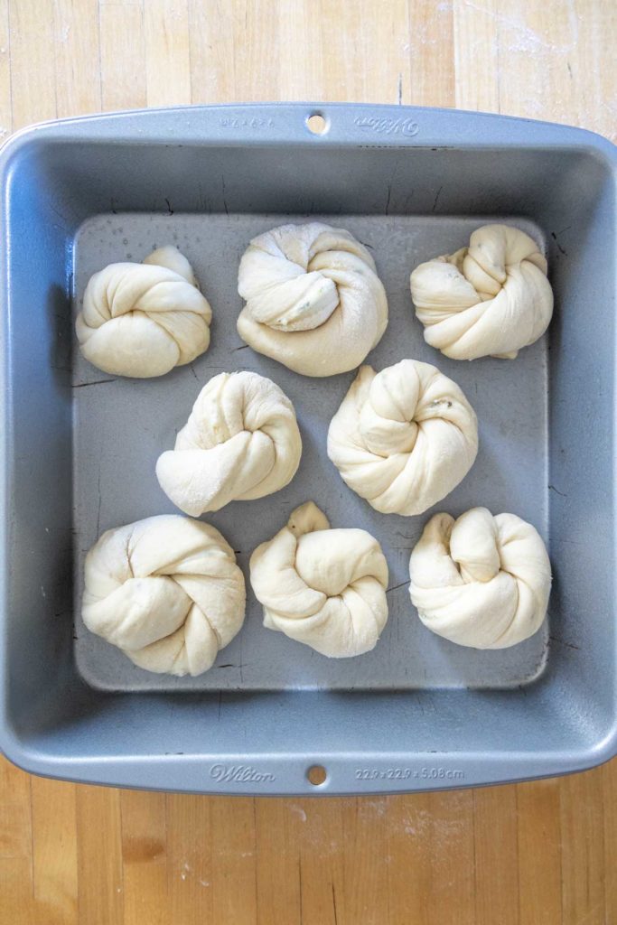 A baking pan with seven unbaked, knot-shaped bread dough pieces arranged inside.