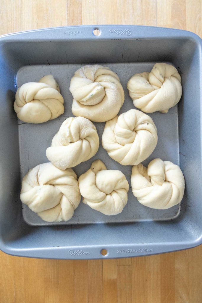 Eight raw, knotted dough pieces in a square baking pan on a wooden surface.