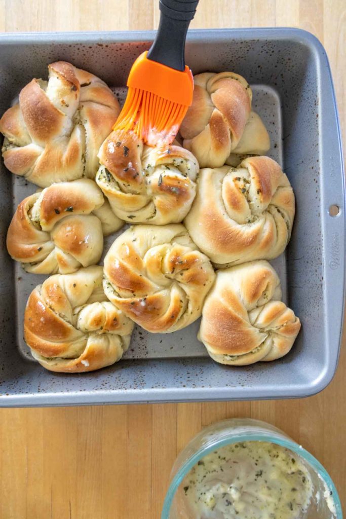 A baking pan with six twisted bread rolls being brushed with melted butter using an orange brush. A bowl of butter and herbs is nearby.