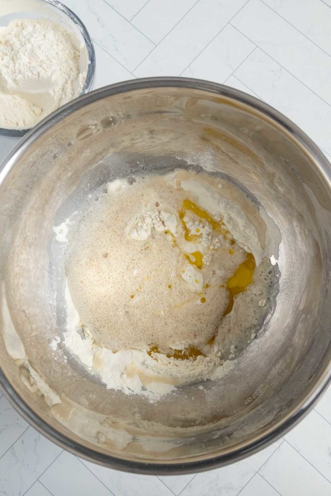 A mixing bowl with flour, liquid, and oil on a white tiled surface; another bowl with flour is partially visible in the background.