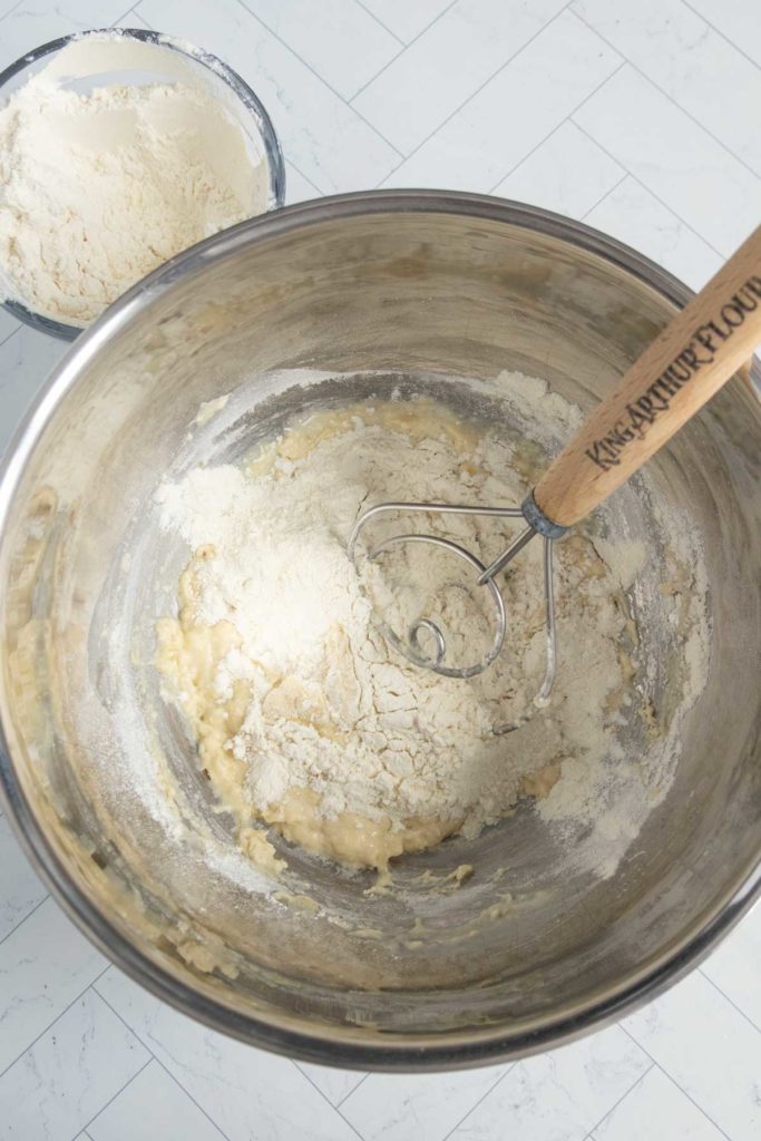 Mixing bowl with dough being stirred by a whisk labeled "King Arthur Flour." A separate bowl with flour is in the background on a tiled kitchen floor.