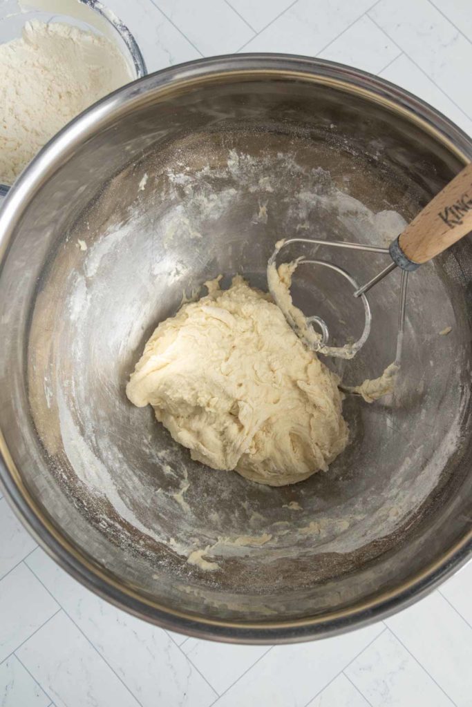 A ball of dough in a metal bowl with a wooden-handled dough hook on a tiled floor.