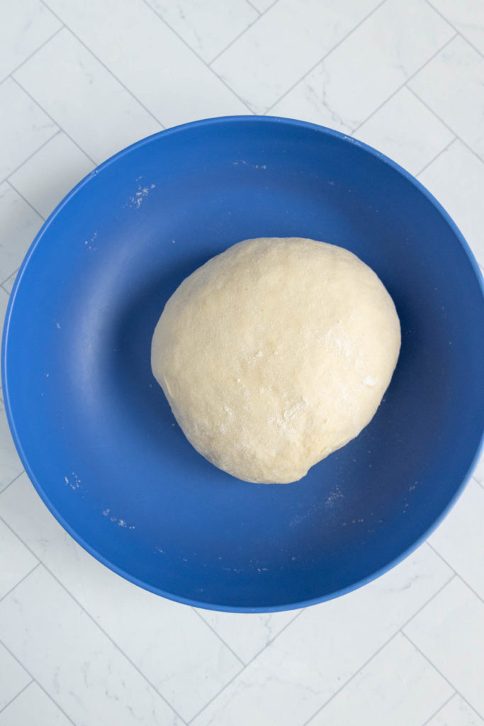 A round dough ball sits in the center of a blue bowl on a tiled surface.