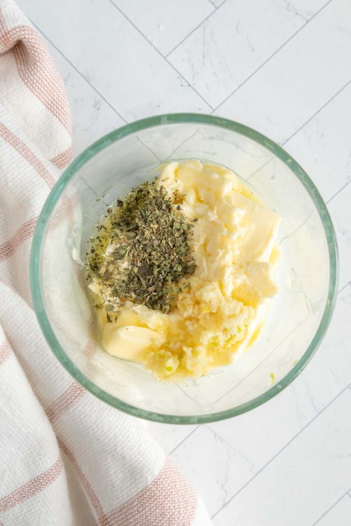 Glass bowl with butter, minced garlic, and dried herbs on a light tiled surface, next to a striped cloth.