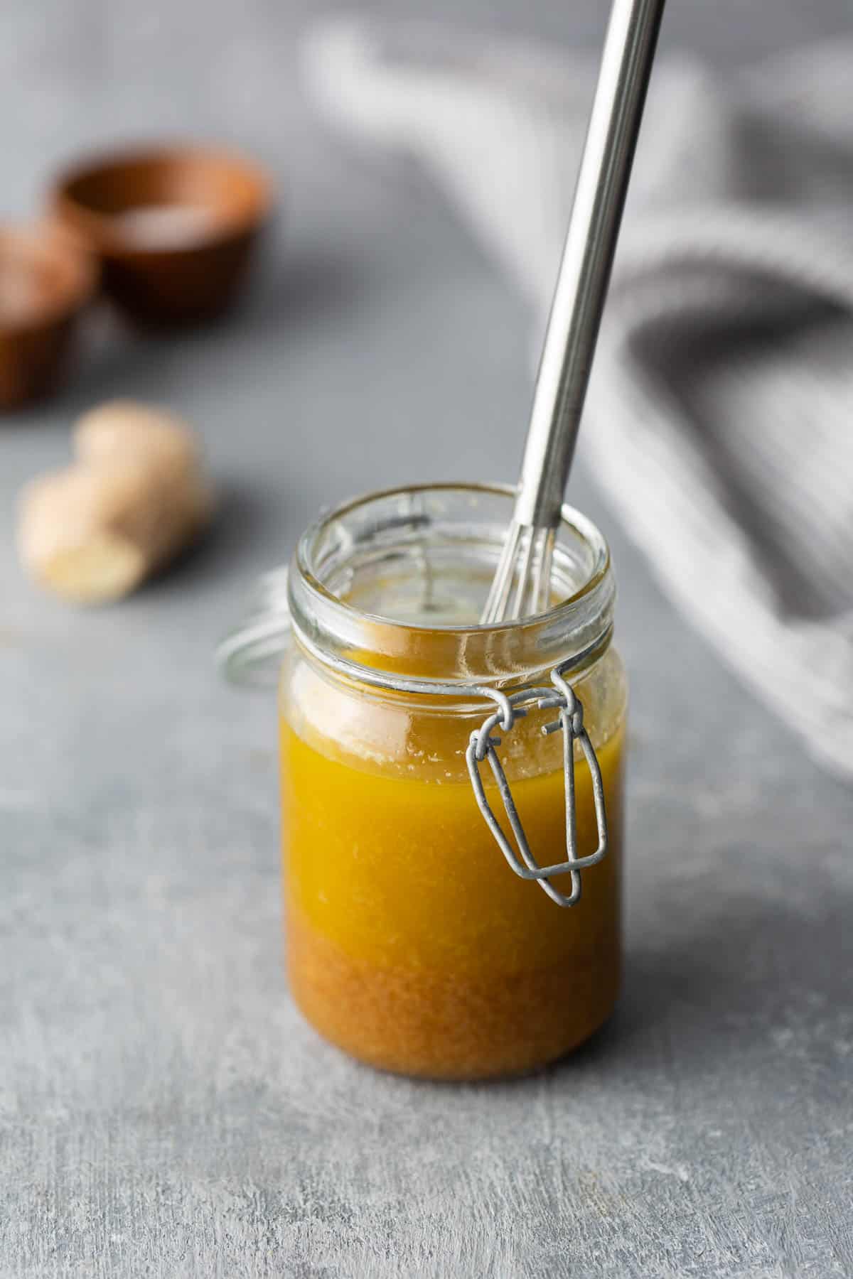 A glass jar filled with yellow liquid and a metal whisk inside sits on a gray surface. Two small bowls and a striped cloth are blurred in the background.