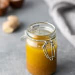 A jar filled with yellow liquid, secured with a metal clip lid, placed on a gray surface. Blurred background includes small wooden bowls and a piece of ginger.