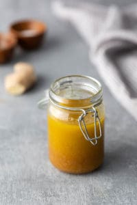 A jar filled with yellow liquid, secured with a metal clip lid, placed on a gray surface. Blurred background includes small wooden bowls and a piece of ginger.