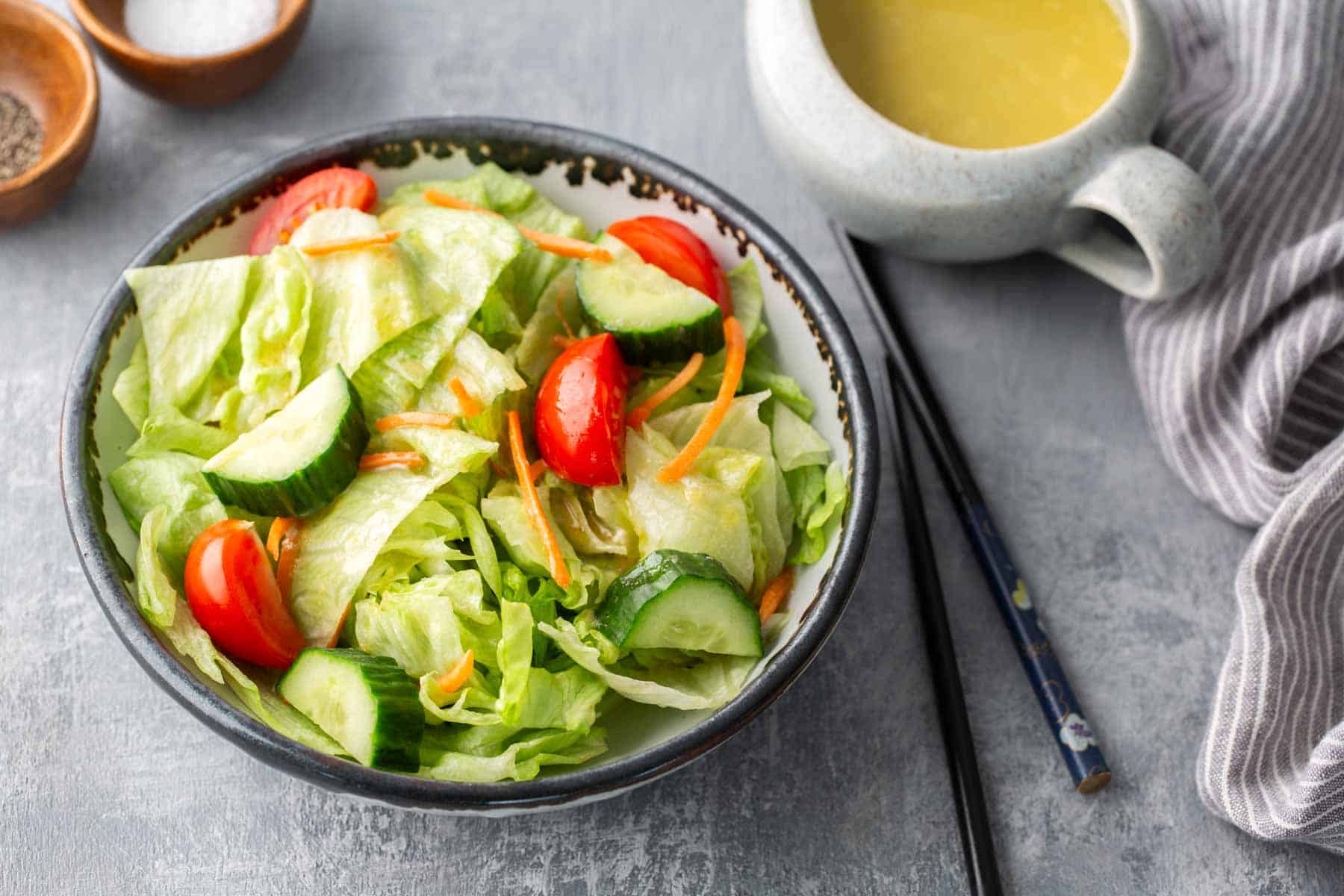 A bowl of mixed salad with lettuce, cucumber, and tomatoes. A cup of dressing is beside it on a gray surface with a striped cloth and chopsticks nearby.