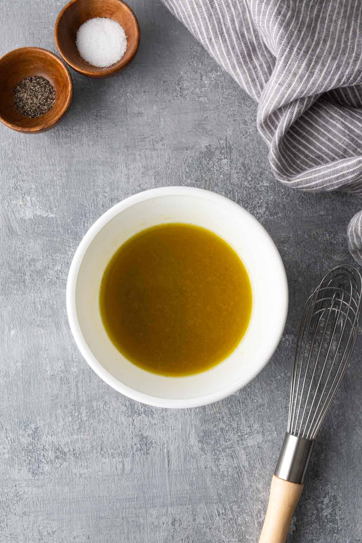 A white bowl of olive oil dressing on a gray surface, next to small bowls of salt and pepper. A stainless steel whisk and striped cloth are nearby.