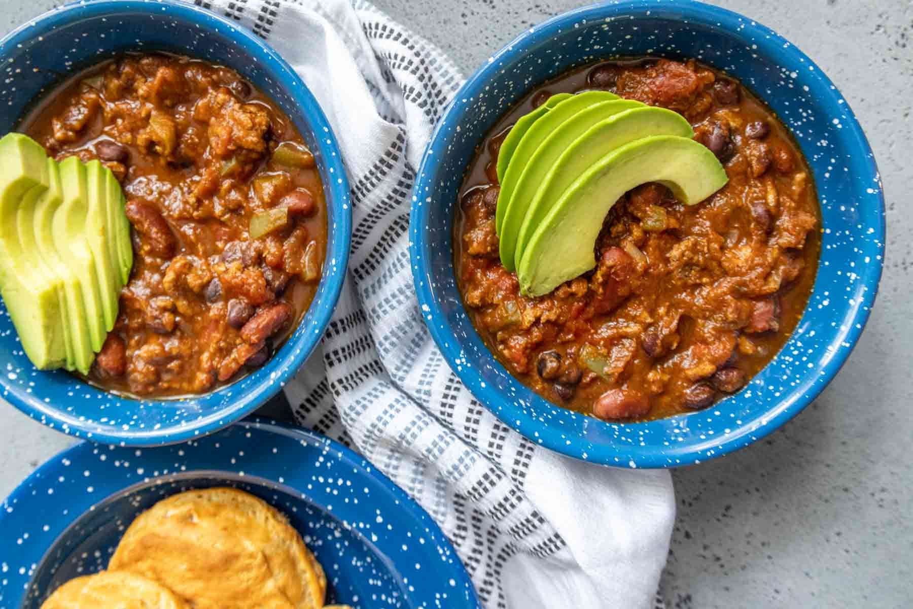 Two blue bowls of chili topped with sliced avocado are set on a table with a white and black striped cloth. A plate of biscuits is visible beside the bowls.