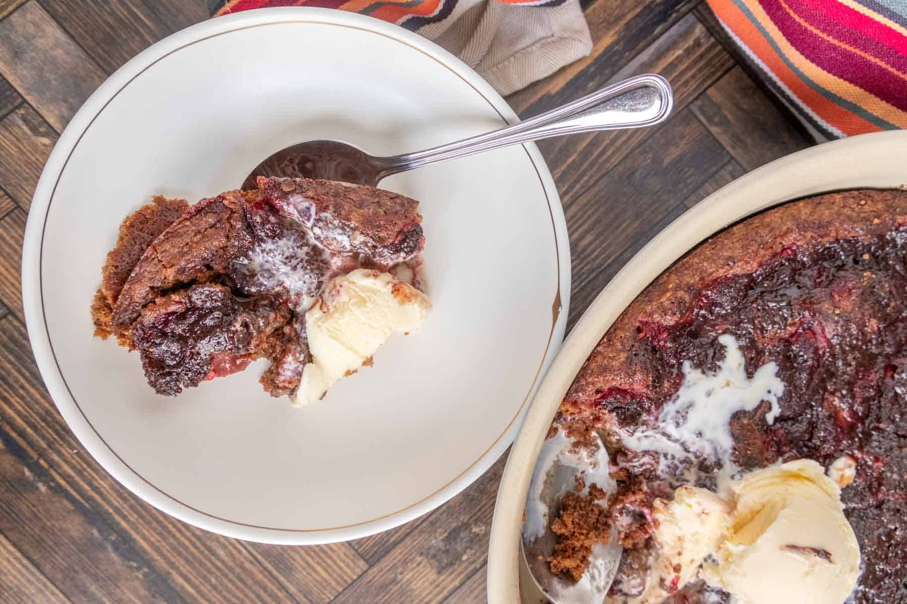 A plate with a serving of chocolate cherry dump cake and vanilla ice cream, alongside a larger dish containing more of the dessert. A spoon rests on the plate.