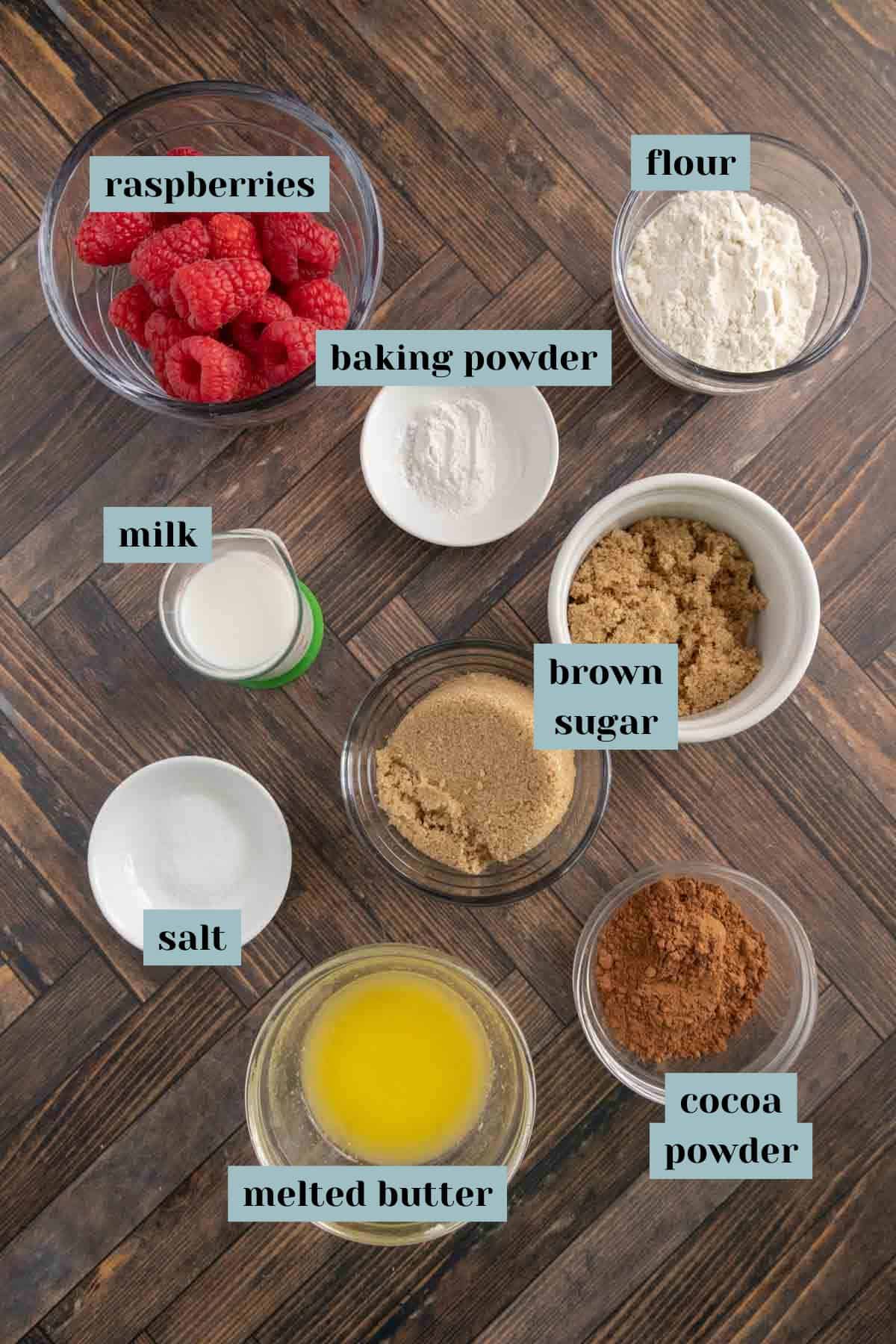 Various labeled ingredients on a wooden surface: raspberries, flour, baking powder, brown sugar, cocoa powder, melted butter, salt, and milk.