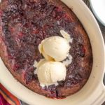 A baked dessert with a dark, fruit-filled surface in an oval dish, topped with two scoops of vanilla ice cream, on a wooden table.