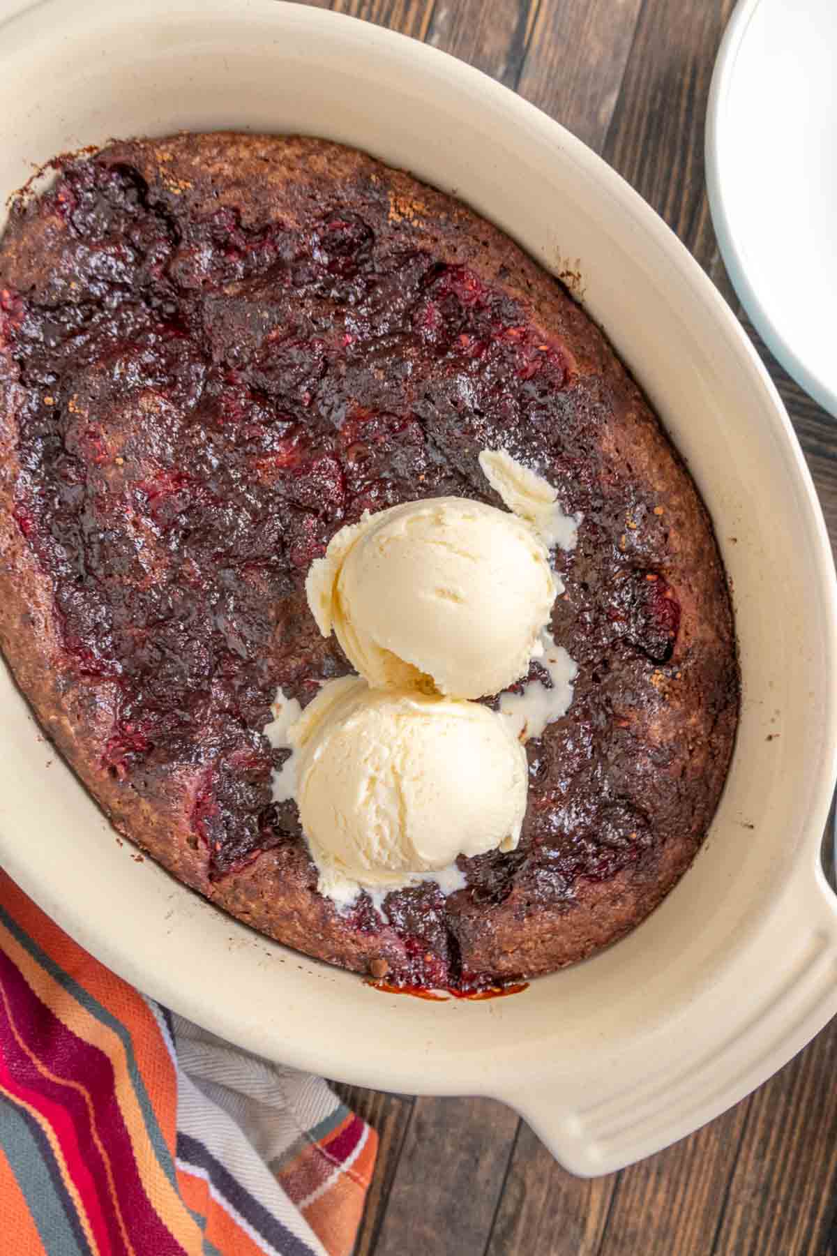 A baked dessert with a dark, fruit-filled surface in an oval dish, topped with two scoops of vanilla ice cream, on a wooden table.