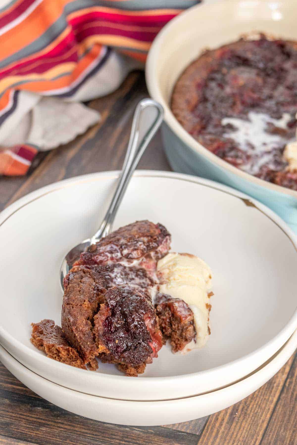 A serving of chocolate cherry cobbler with a scoop of vanilla ice cream on a white plate, with a spoon beside it. A multicolor striped cloth is seen in the background.