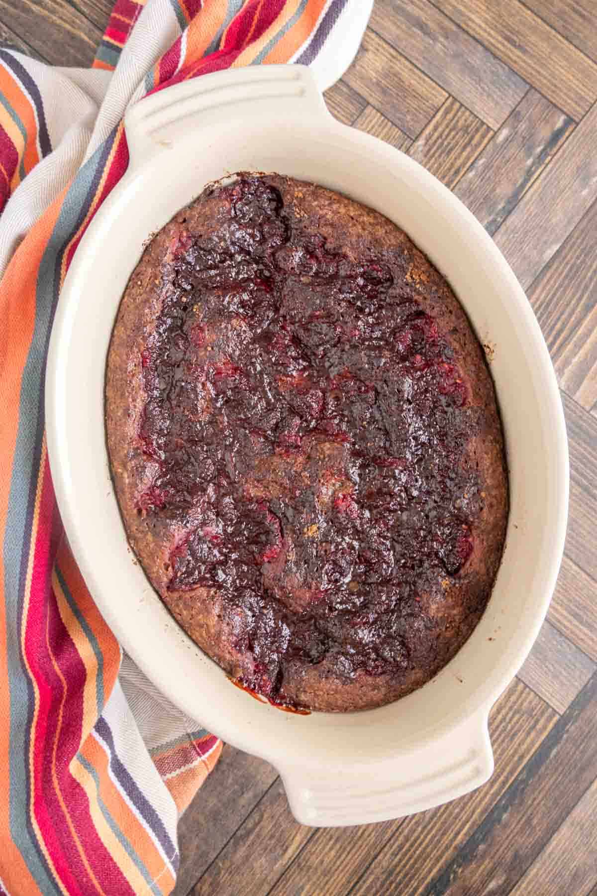 Baked dessert in an oval dish with a striped cloth beside it, featuring a dark, textured surface indicative of fruit or berry topping.