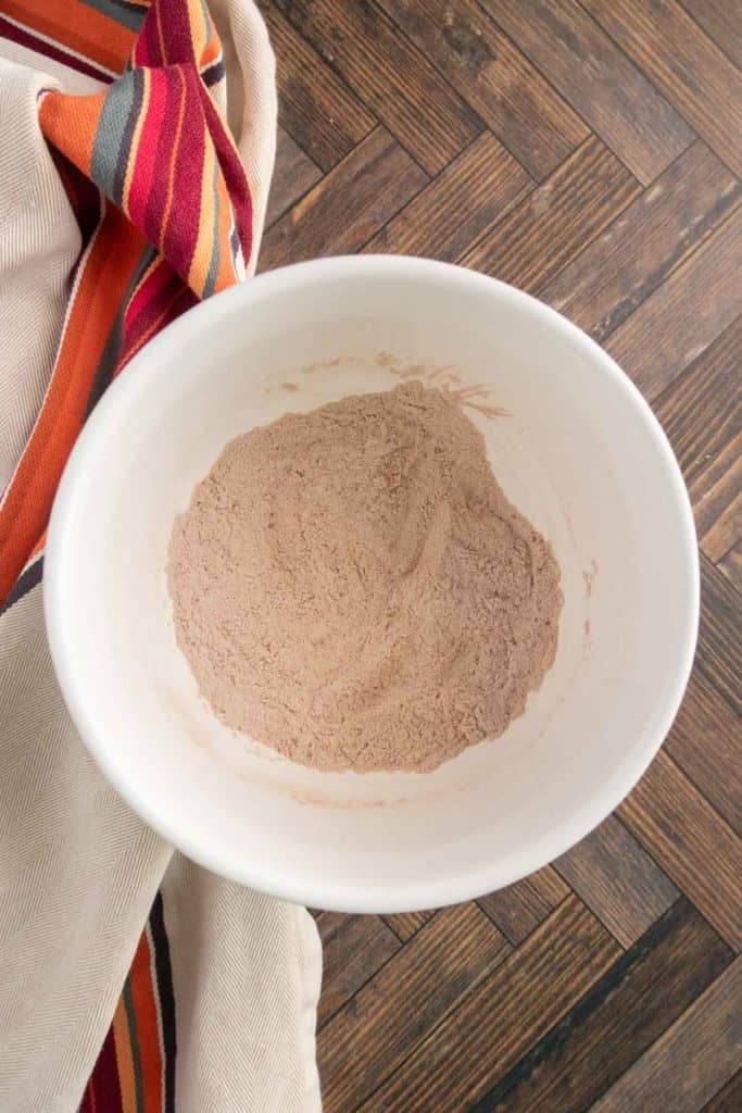 A white bowl containing a mound of cocoa powder on a wooden surface with a striped cloth nearby.