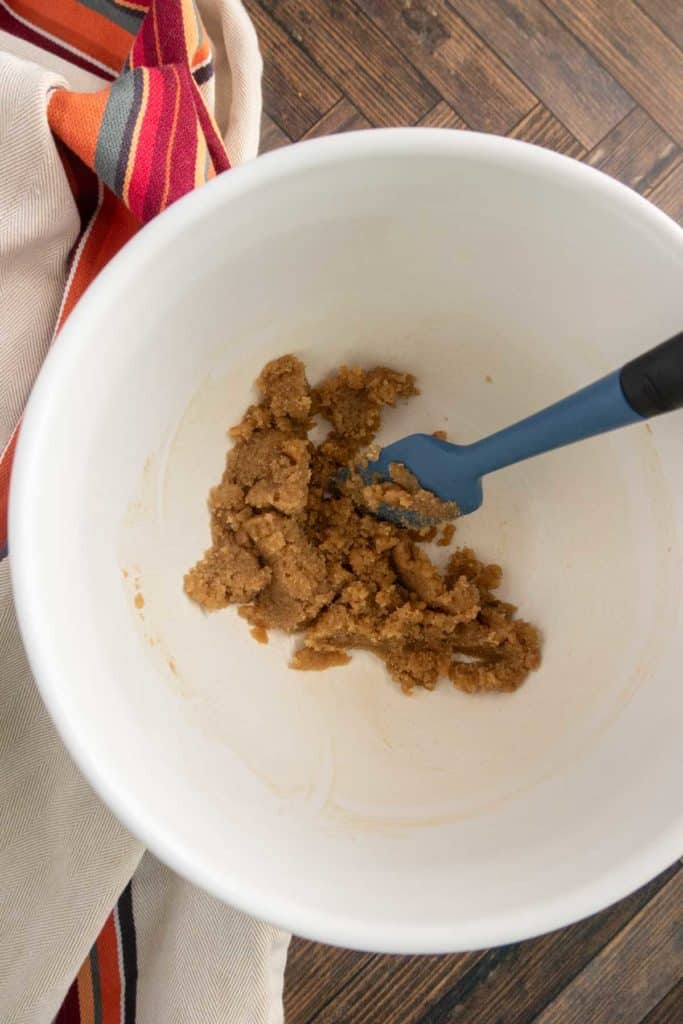 A blue spatula rests in a white bowl containing brown sugar on a wooden surface, next to a striped cloth.