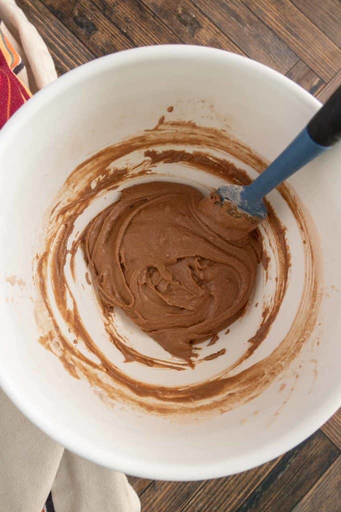 Chocolate batter in a white mixing bowl with a blue spatula on a wooden surface.