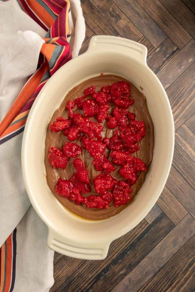 Ceramic dish with chocolate batter topped with raspberries, placed on a wooden surface. A striped cloth lies beside the dish.