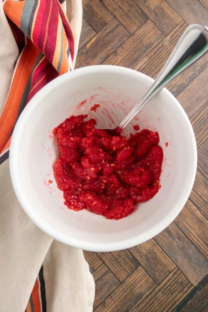 A white bowl with mashed raspberries and a spoon inside, on a wooden surface alongside a striped cloth.