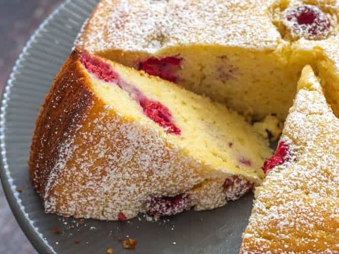 close up of sliced ricotta cake with raspberries and lemon on a gray cake plate