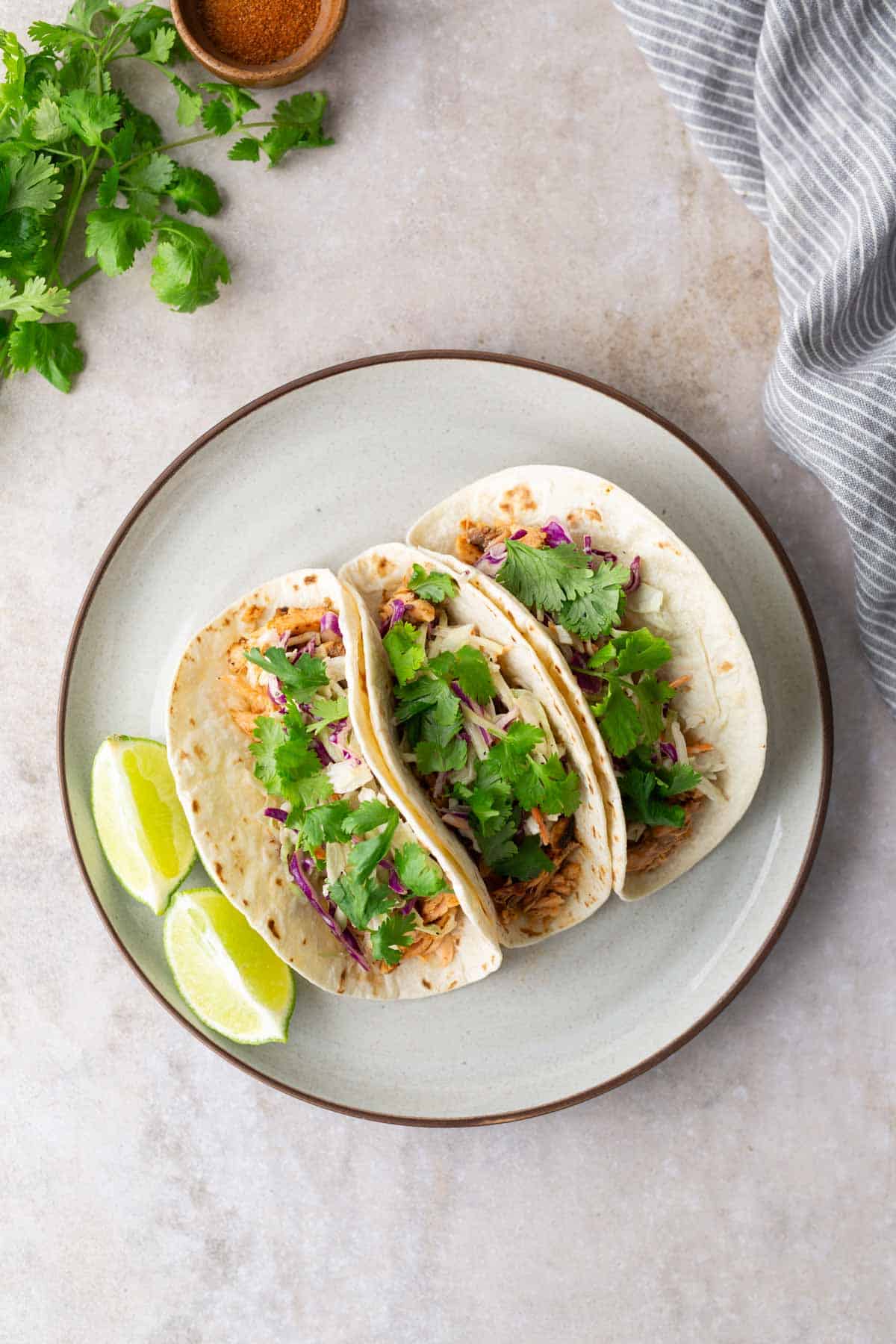 Three tacos filled with shredded meat, cabbage, and cilantro on a plate with lime wedges and a striped cloth nearby.