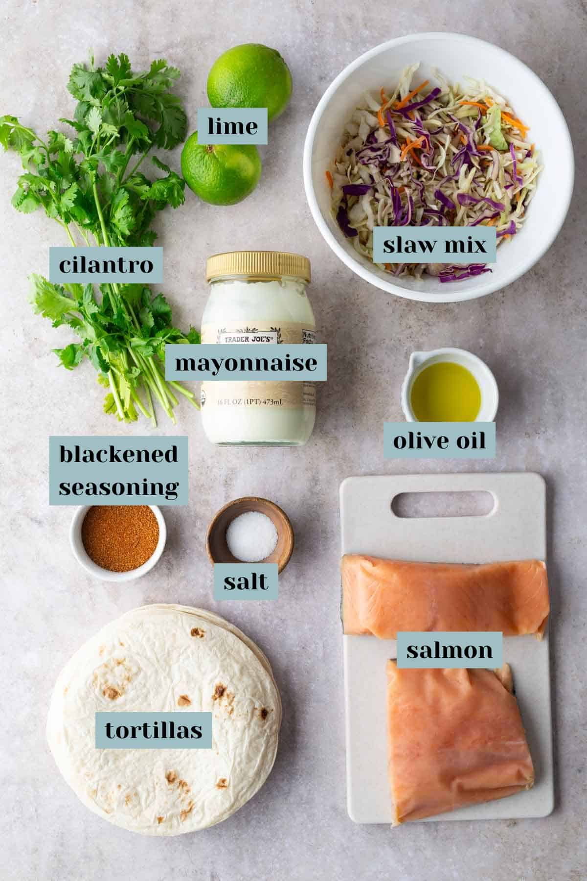 Ingredients for a salmon wrap: lime, cilantro, slaw mix, mayonnaise, olive oil, blackened seasoning, salt, tortillas, and salmon fillets arranged on a light surface.