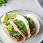 Two tacos filled with shredded chicken, cabbage, and cilantro on a plate. Lime wedges are placed beside the tacos.