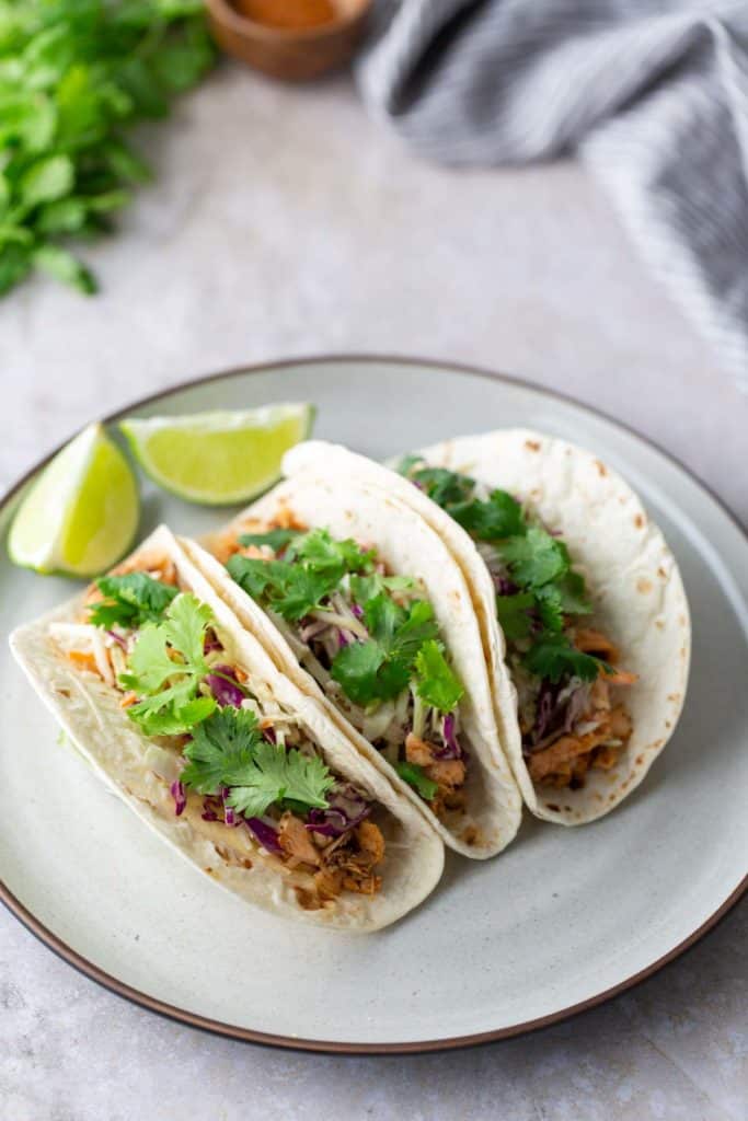 Two tacos filled with shredded chicken, cabbage, and cilantro on a plate. Lime wedges are placed beside the tacos.