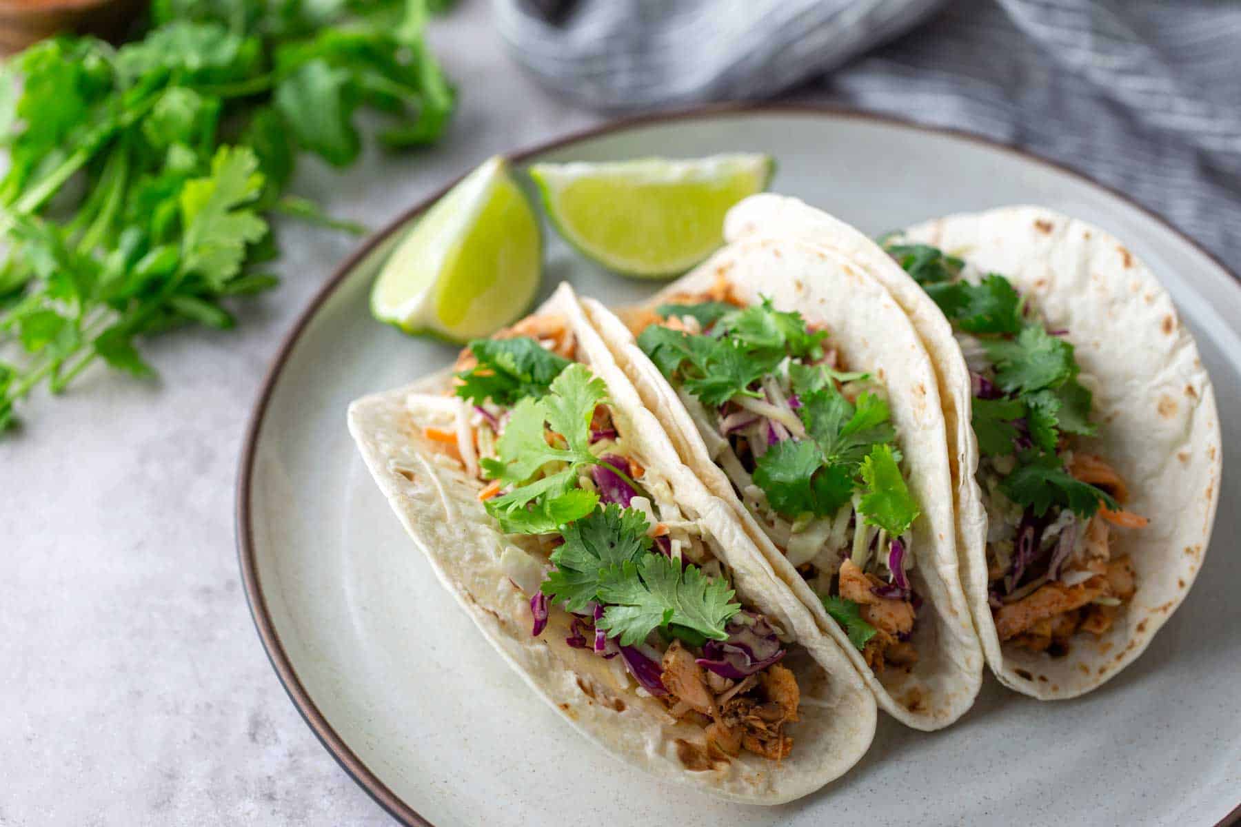 Three soft tacos filled with meat, cabbage, and cilantro on a plate, with lime wedges and cilantro in the background.