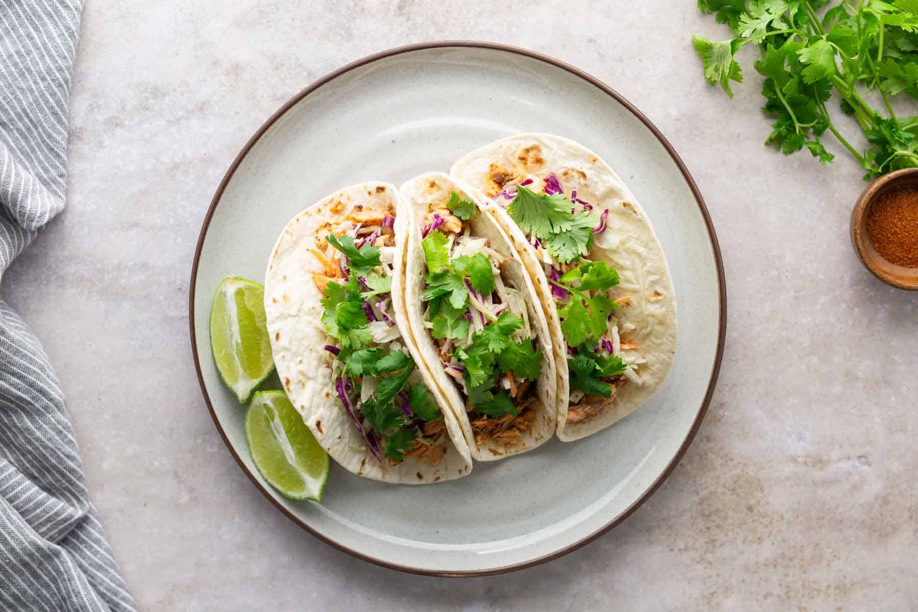 Three tacos with cilantro and shredded filling on a plate, accompanied by lime wedges and garnished with herbs.