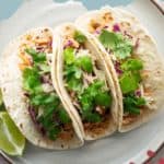 Plate of three salmon tacos, garnished with cilantro and cabbage slaw, with lime wedges on the side.