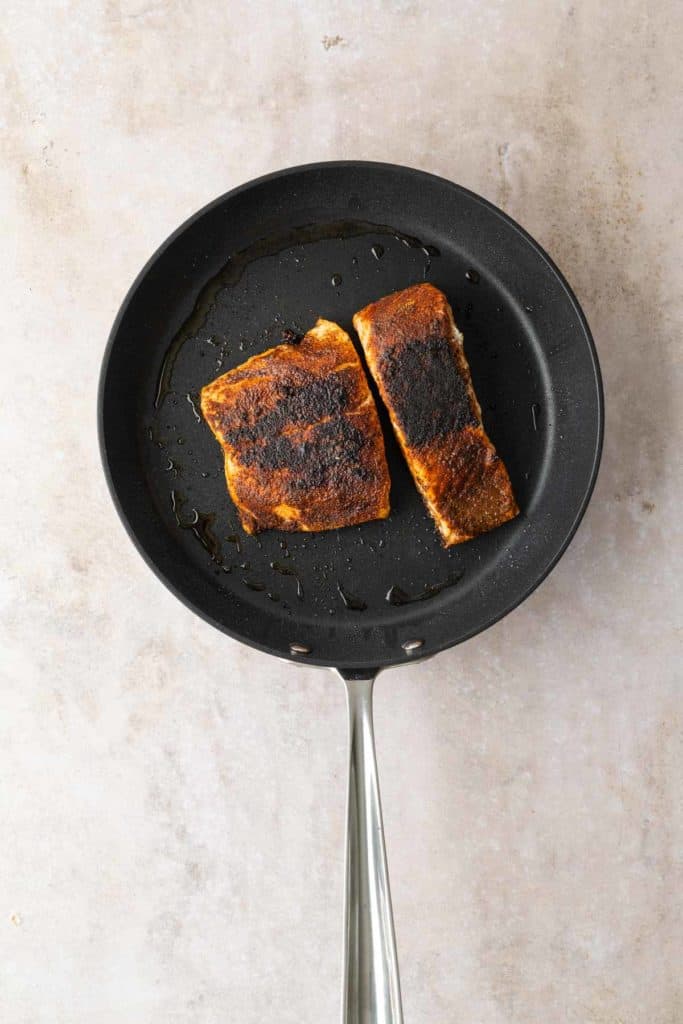 Two pieces of cooked salmon with charred seasoning in a black frying pan.