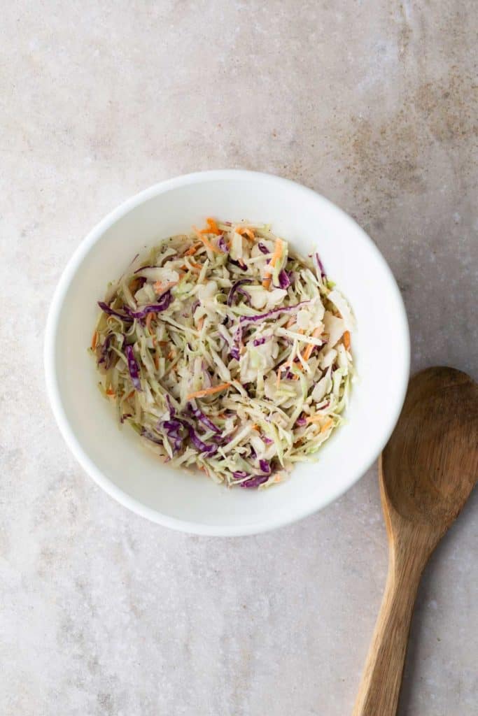 A bowl of coleslaw with shredded cabbage and carrots on a textured surface, accompanied by a wooden spoon.