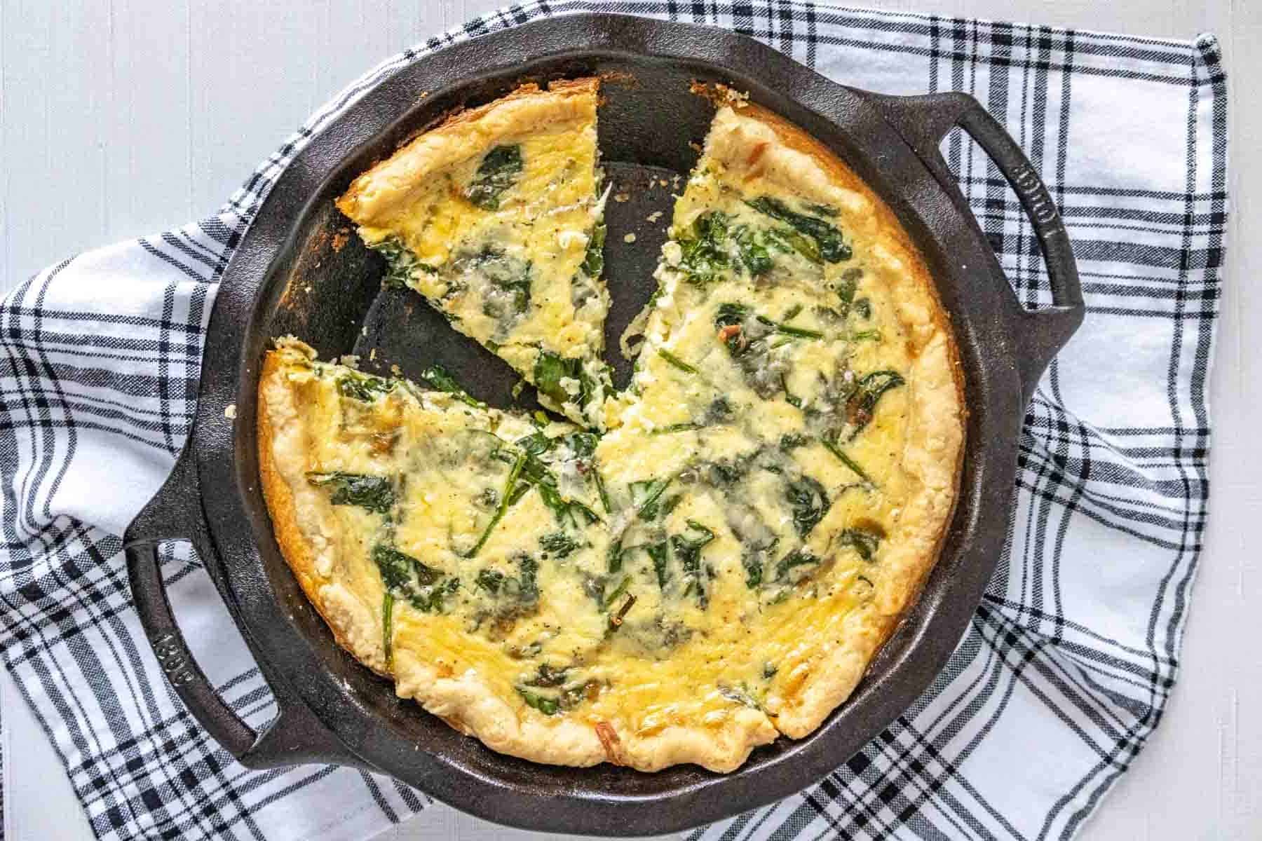 Spinach quiche in a cast iron pie plate with a black and white kitchen towel.