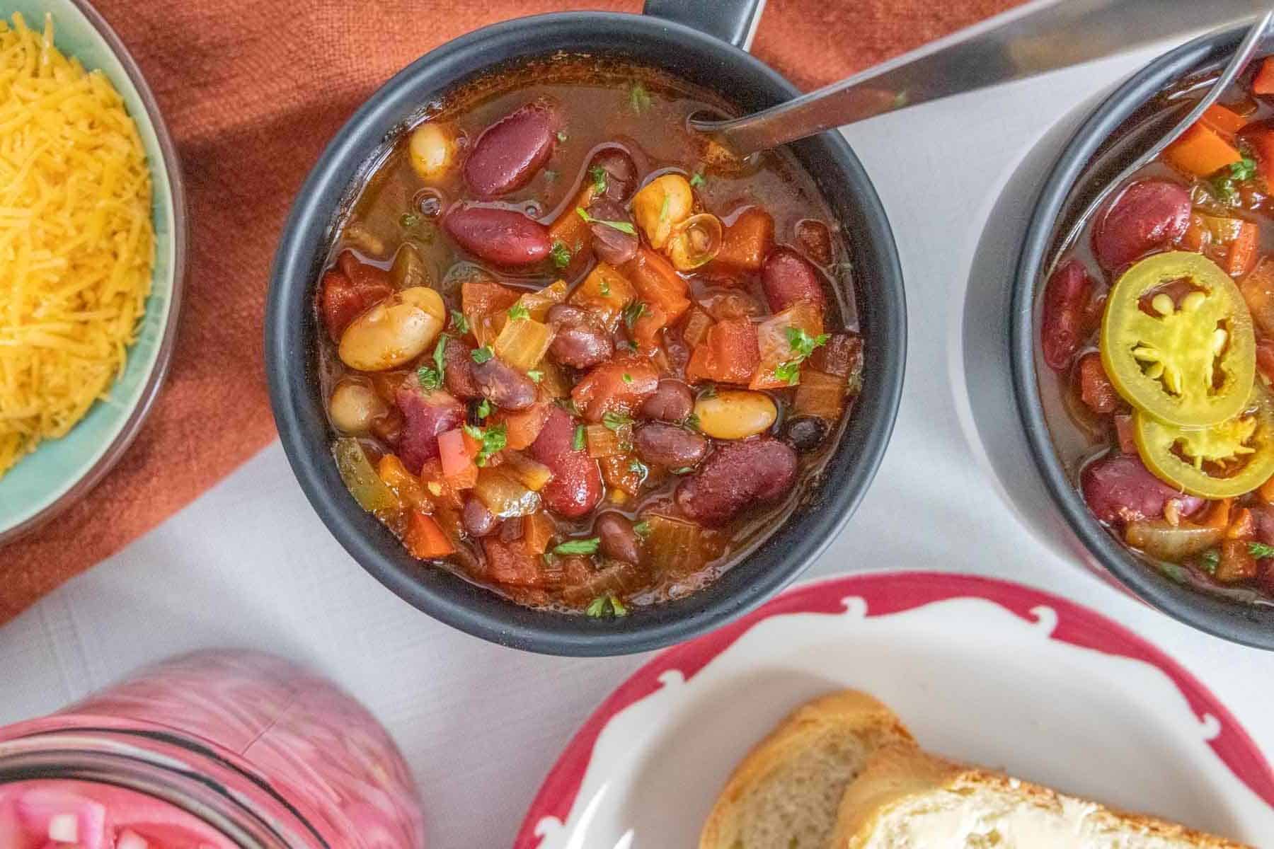 Overhead of mugs of three bean chili with bread, cheese, and pickled onions off the sides.