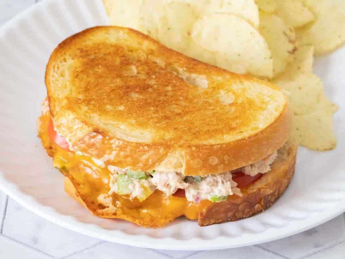 Tuna melt sandwich on white plate with potato chips beside.