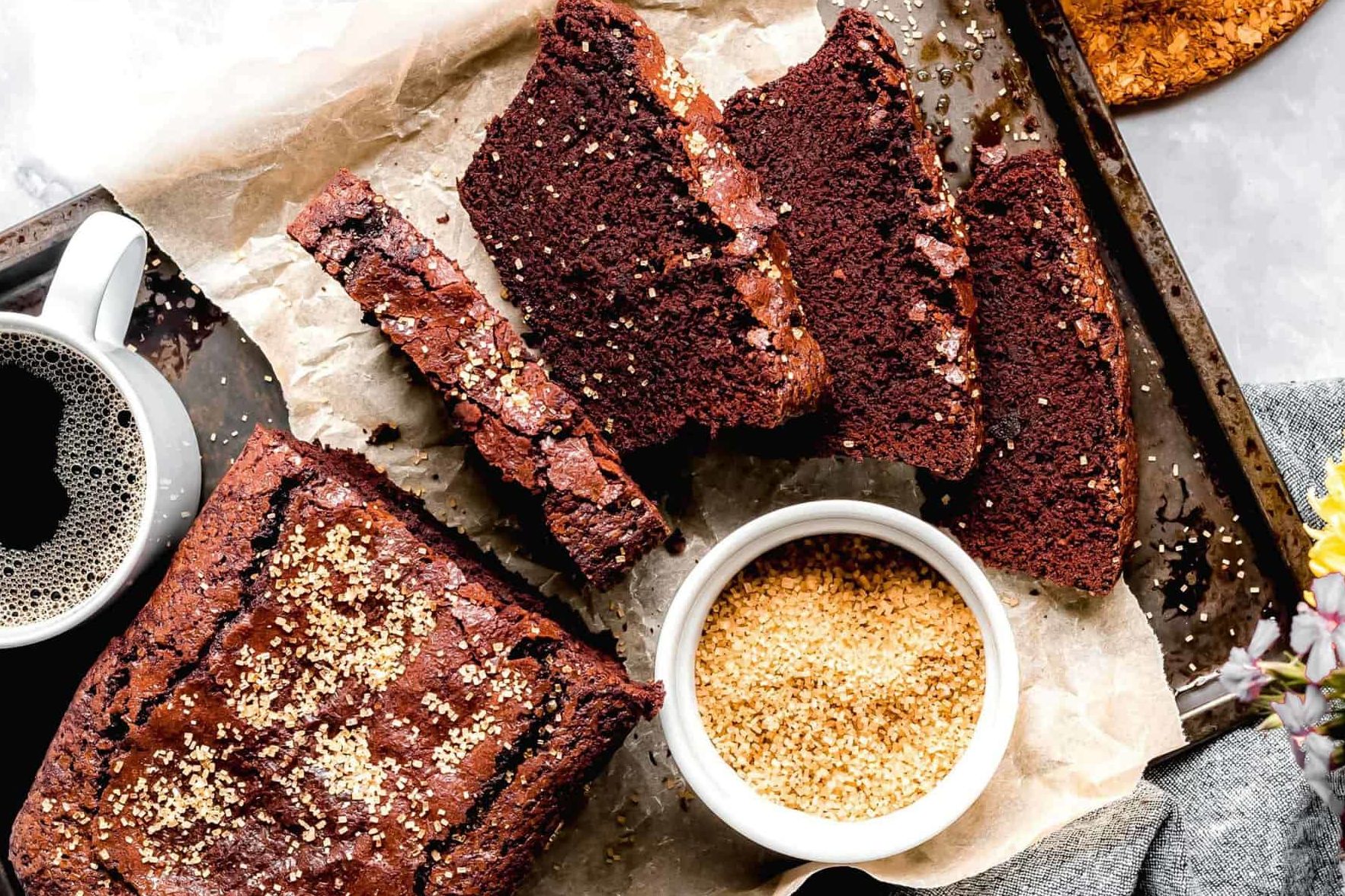 A tray with sliced chocolate bread, a cup of coffee, and a small bowl of sesame seeds on parchment paper.