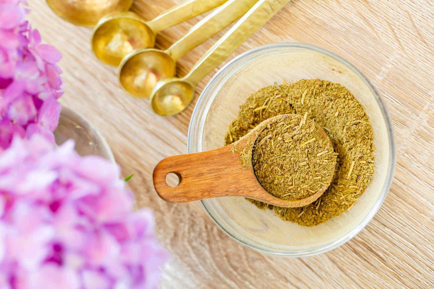 A clear bowl with a wooden spoon contains herbal powder. Golden measuring spoons are beside it. Pink flowers are blurred in the background.