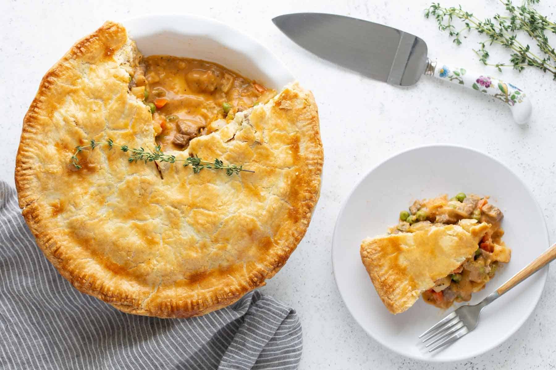 Beef pot pie with a slice served on a white plate, accompanied by a knife and fresh thyme sprigs on a marble surface.