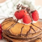 A stack of pancakes topped with whipped cream, raspberries, and chocolate sauce on a plate.