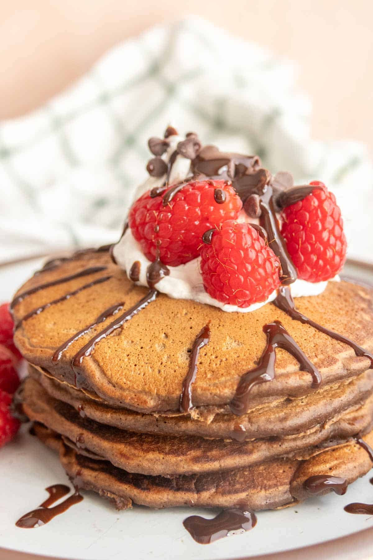 A stack of pancakes topped with whipped cream, raspberries, and chocolate sauce on a plate.