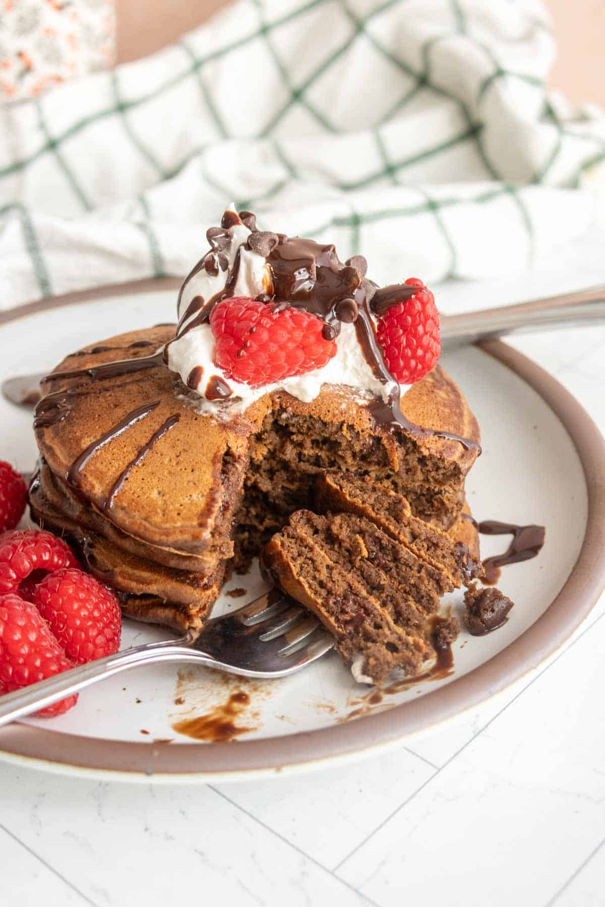 A stack of chocolate pancakes topped with whipped cream, raspberries, and chocolate drizzle on a plate with a fork. Some pancakes are cut, and extra raspberries are on the side.