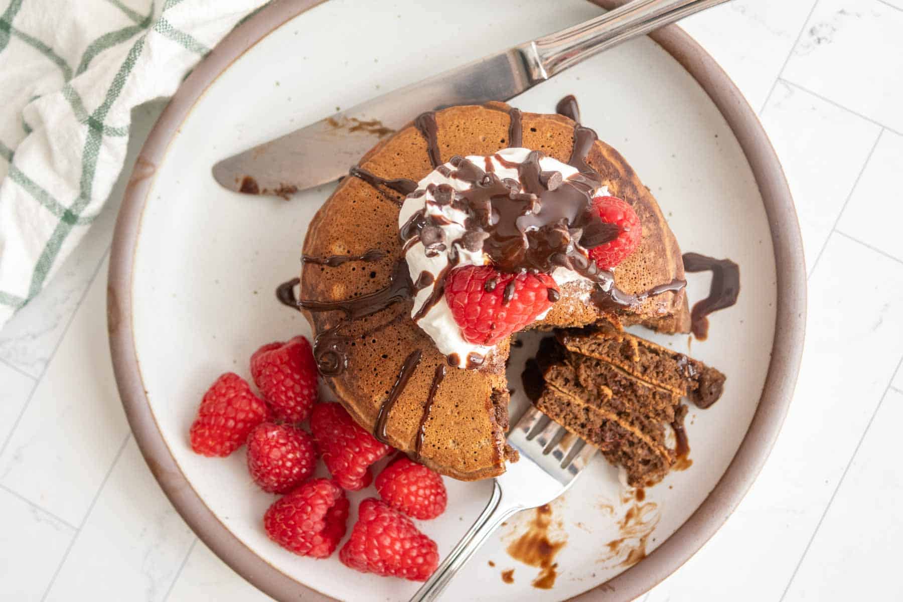 Mini bundt cake with chocolate drizzle, whipped cream, and raspberries on a plate. Fork with cake slices and a knife beside it.