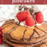 Stack of chocolate pancakes topped with whipped cream, fresh raspberries, and chocolate drizzle on a plate.