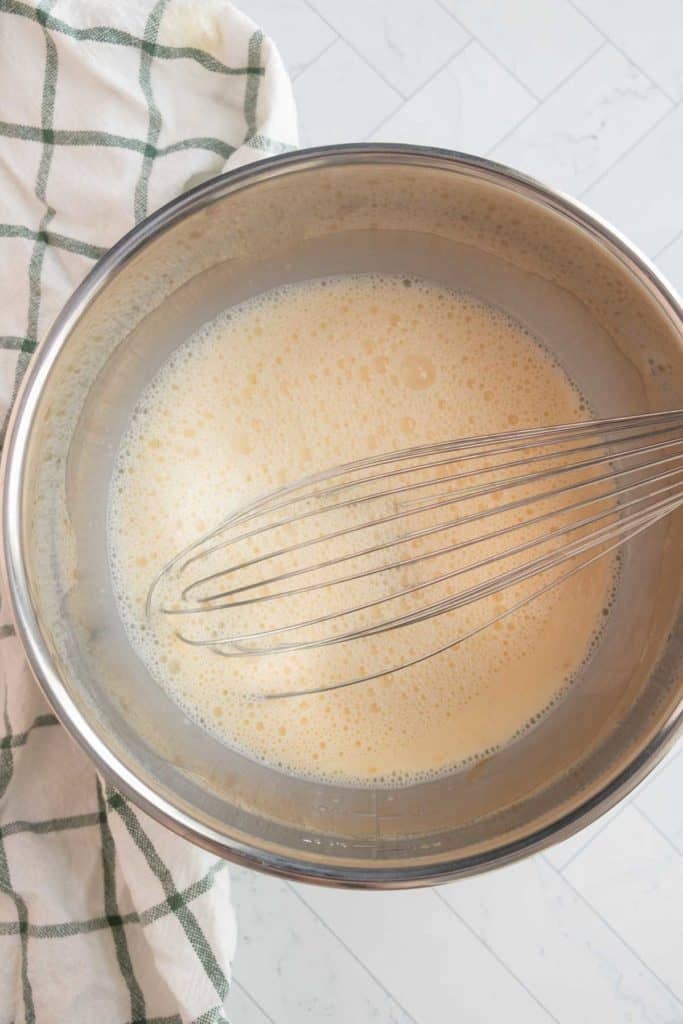 A whisk in a metal bowl containing a frothy liquid mixture, placed on a white surface with a green and white checkered cloth beside it.